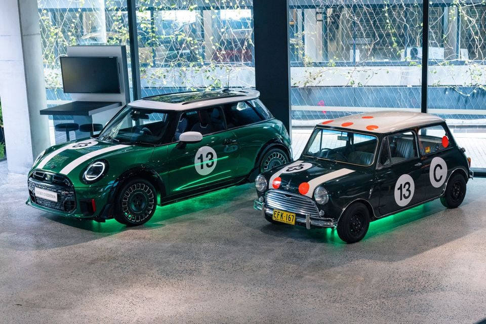 A green Mini Cooper Lucky 13 Edition hatch beside a classic Mini Cooper in matching race livery with number 13, displayed indoors with green underglow