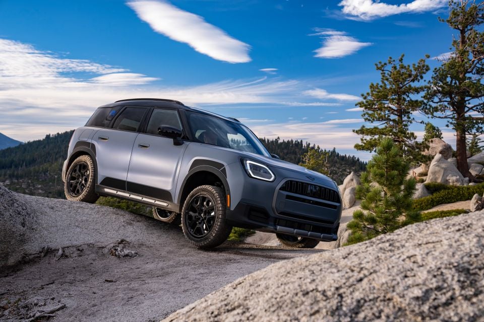 A grey Mini Countryman Untamed Edition SUV parked on rocky terrain with trees and mountains in the background.