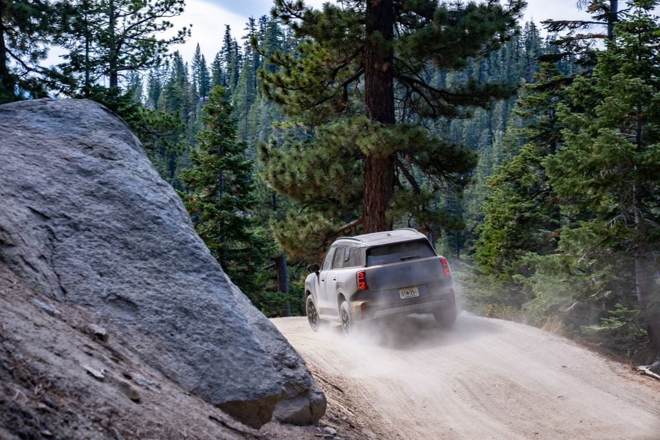 A grey Mini Countryman SUV kicks up dust while driving away from the camera on a forest dirt road