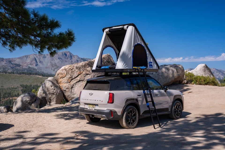 A white Mini Countryman S SUV parked outdoors with a rooftop tent set up, seen from the rear quarter angle.