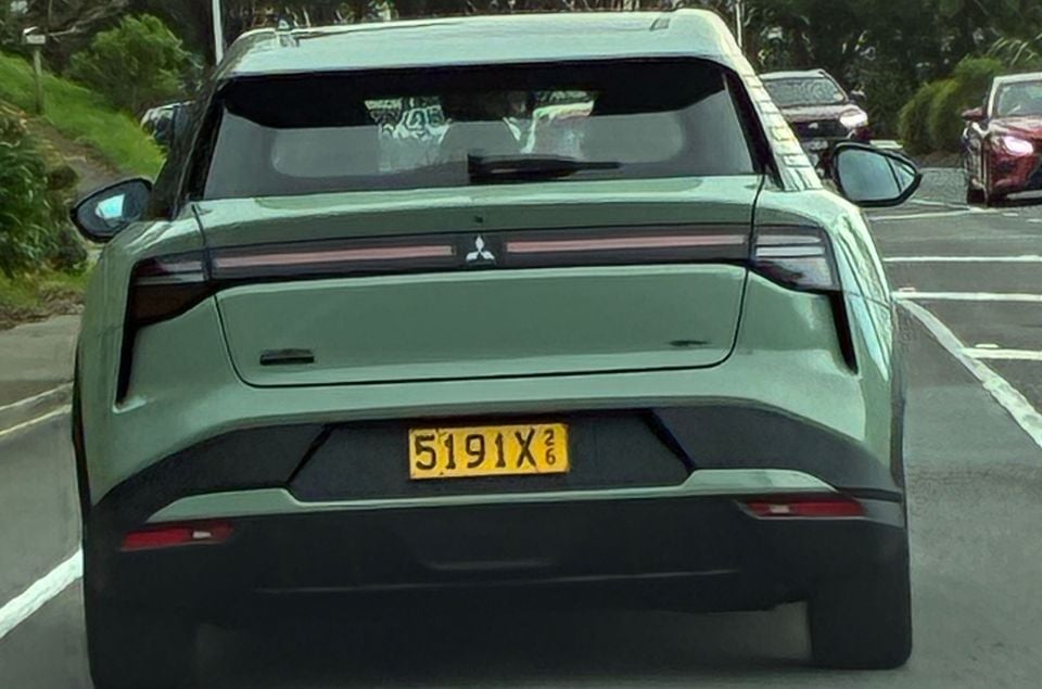Rear view of a green Mitsubishi ASX VR-E electric SUV driving on a road, showing its full-width tail light bar and yellow New Zealand number plate