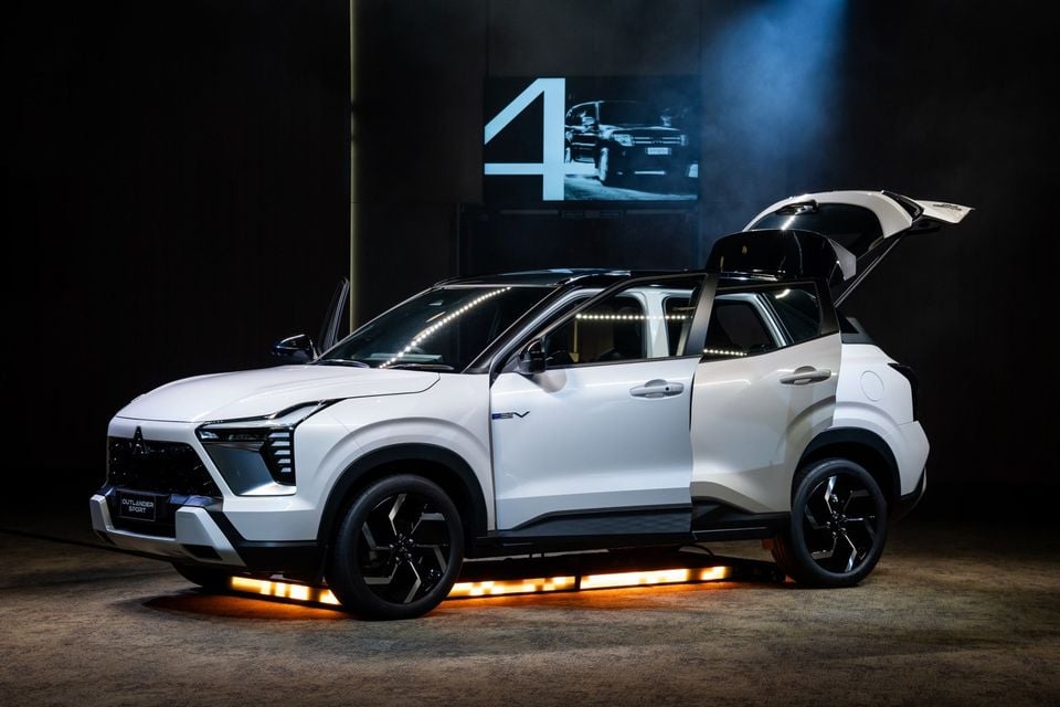 A white Mitsubishi Outlander Sport SUV displayed at an indoor reveal event, shown from the front-side angle with doors open and orange underlighting