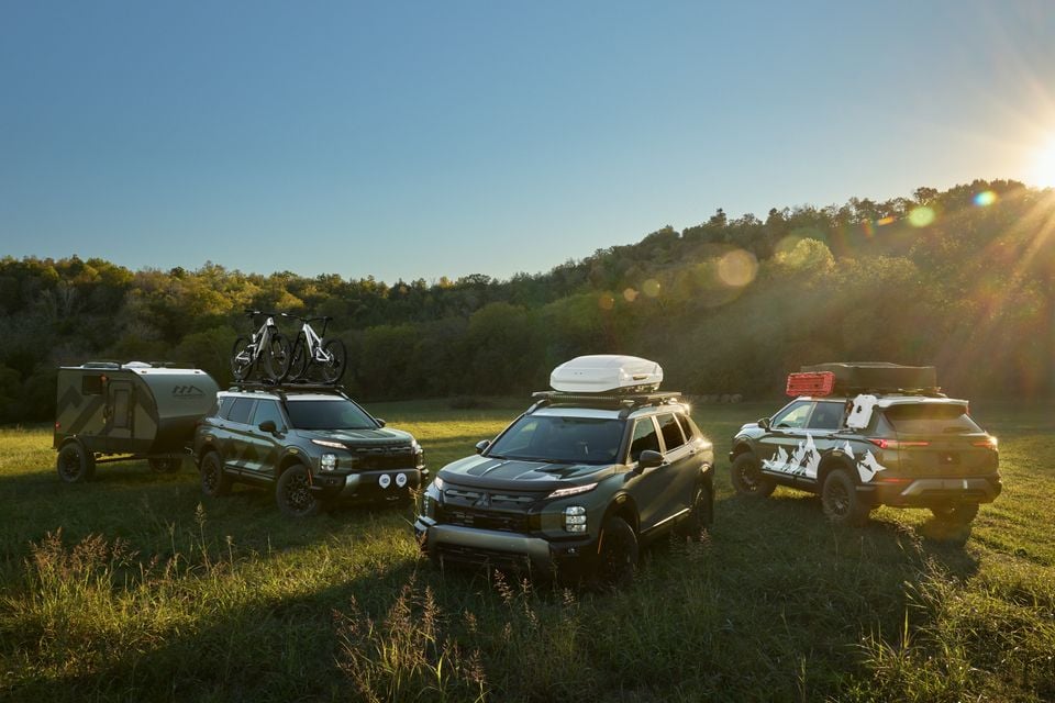 Three customised Mitsubishi Outlander SUVs with roof racks, bikes and a cargo box parked in a grassy field at sunset, one hitched to a camper trailer