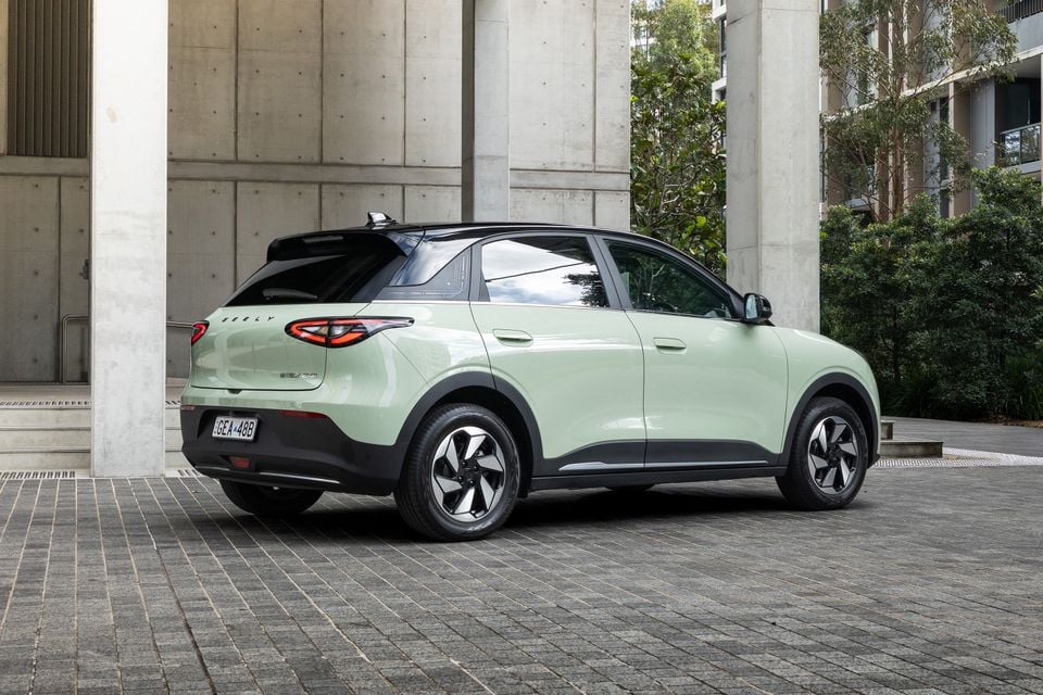 A light green Geely EX2 SUV seen from the rear three-quarter angle, parked on a brick paved area near concrete columns