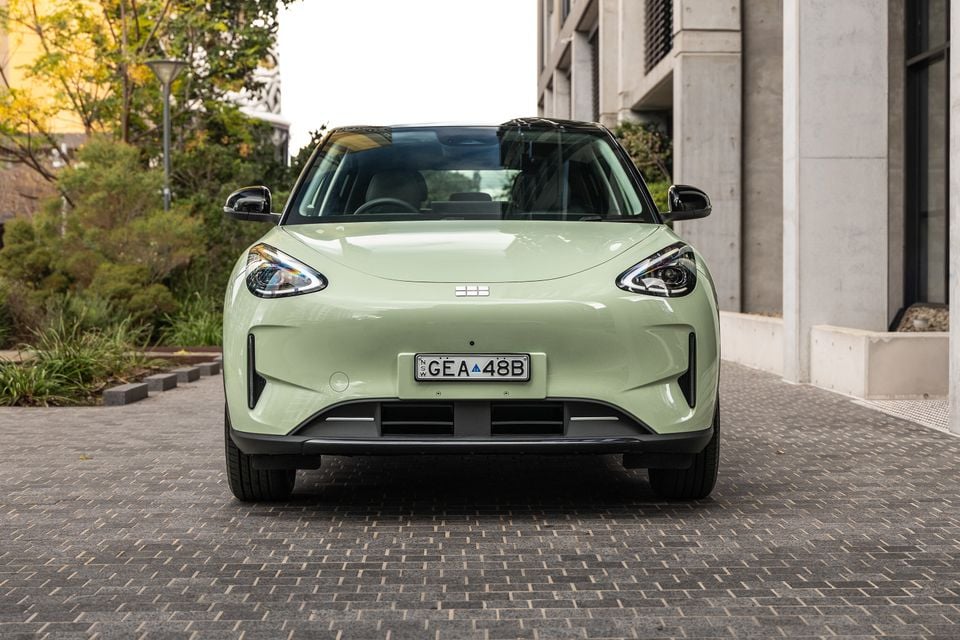 A light green Geely EX2 small SUV seen from the front, parked on a cobblestone surface in an urban setting.