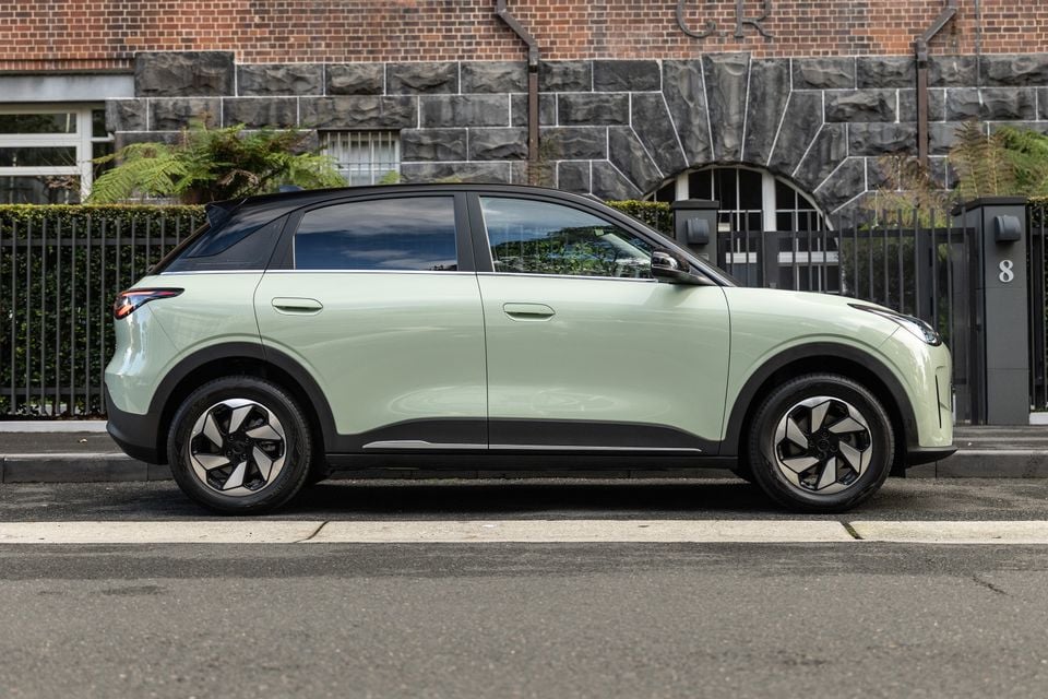 A light green Geely EX2 small SUV seen from the side, parked on a street in front of a brick building.