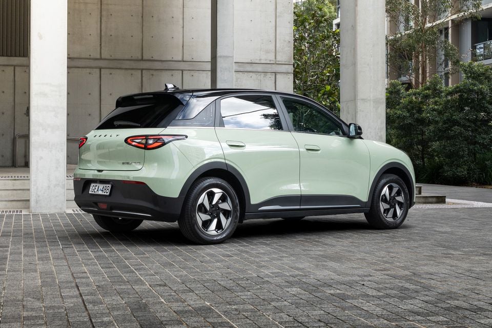 A light green Geely EX2 SUV seen from the rear three-quarter angle, parked on a paved urban area next to concrete columns and trees.