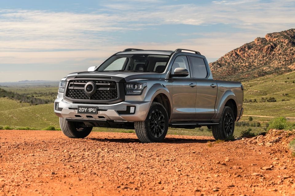 A grey GWM Cannon Alpha dual-cab ute seen from the front-side, parked on a red dirt track with rocky hills and green landscape in the background