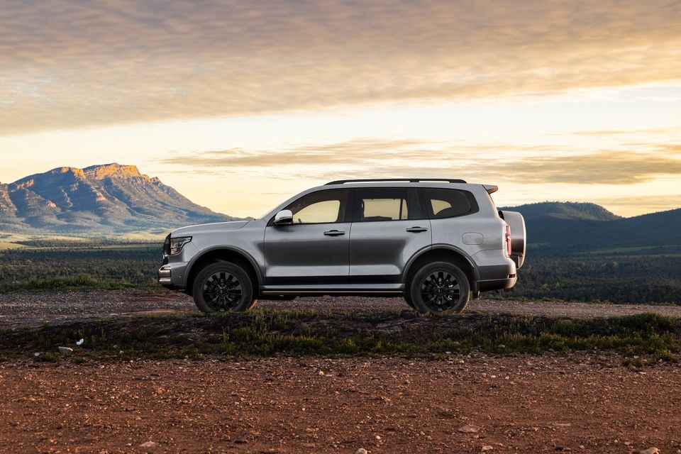 A silver GWM Tank 500 large SUV seen from the side, parked on a rocky hilltop with a mountain landscape and sunset sky in the background.