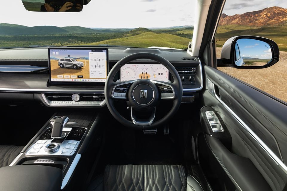 Interior of a GWM Tank 500 showing the dashboard, large touchscreen infotainment display, digital instrument cluster, steering wheel, and centre console, with a scenic hilly landscape visible through the windscreen