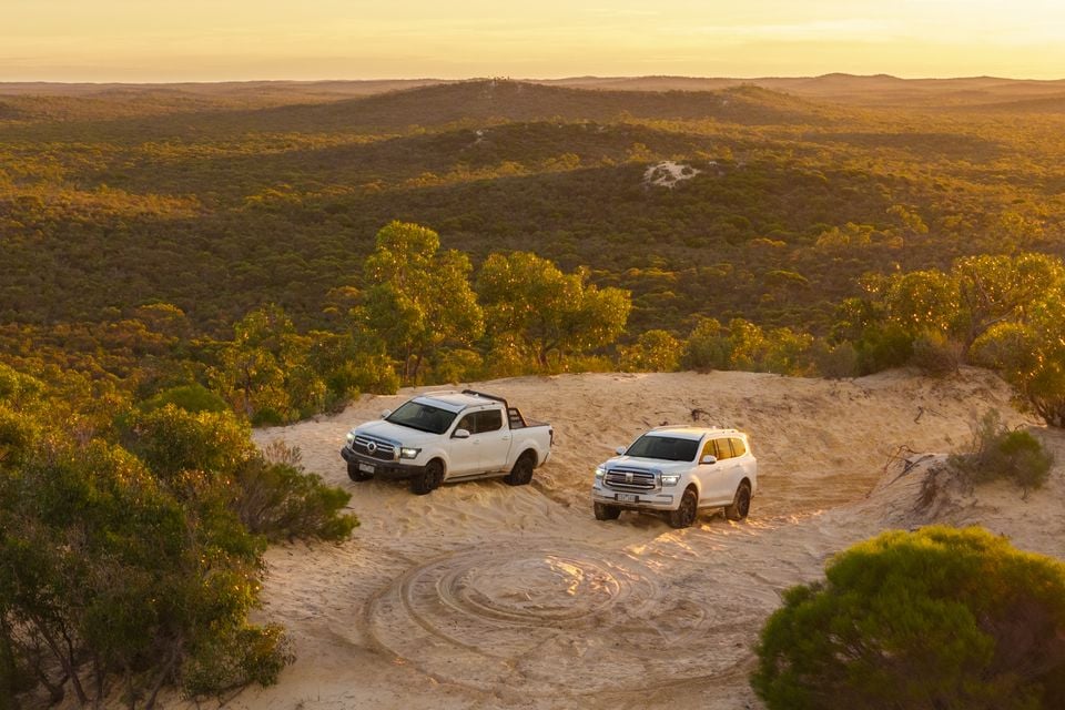 Two white GWM ute and SUV 4WDs parked on a sandy clearing at sunset, surrounded by Australian bushland viewed from above
