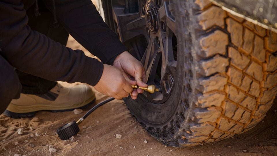 A person connecting an air hose to the tyre valve of a mud-terrain tyre on an off-road vehicle, crouching on red dirt terrain
