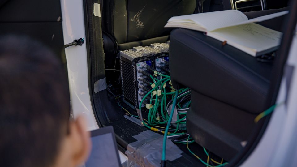 Testing equipment and wiring harnesses installed in the rear footwell of a white SUV, with a person and notebook visible nearby