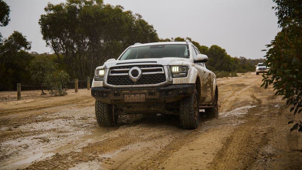 A white GWM Tank 500 ute with a bull bar driving along a muddy dirt track in wet conditions, seen from the front