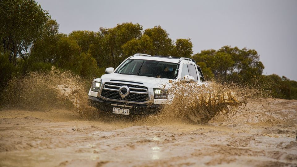 A white GWM Tank 500 SUV powering through a muddy water crossing on an outback dirt track, seen from the front