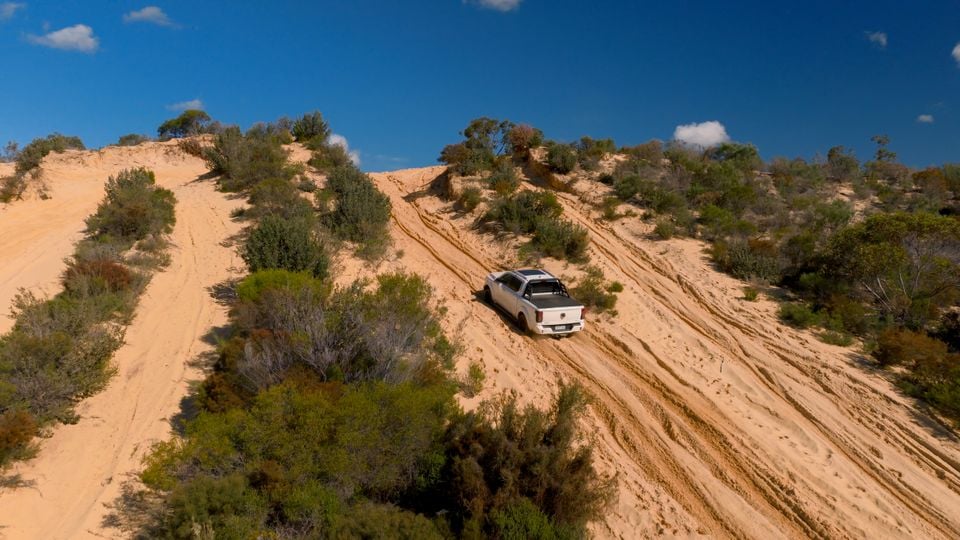 A white GWM Tank 500 ute climbing a steep sandy track on an off-road trail, seen from a drone aerial perspective with scrubland and blue sky in the background