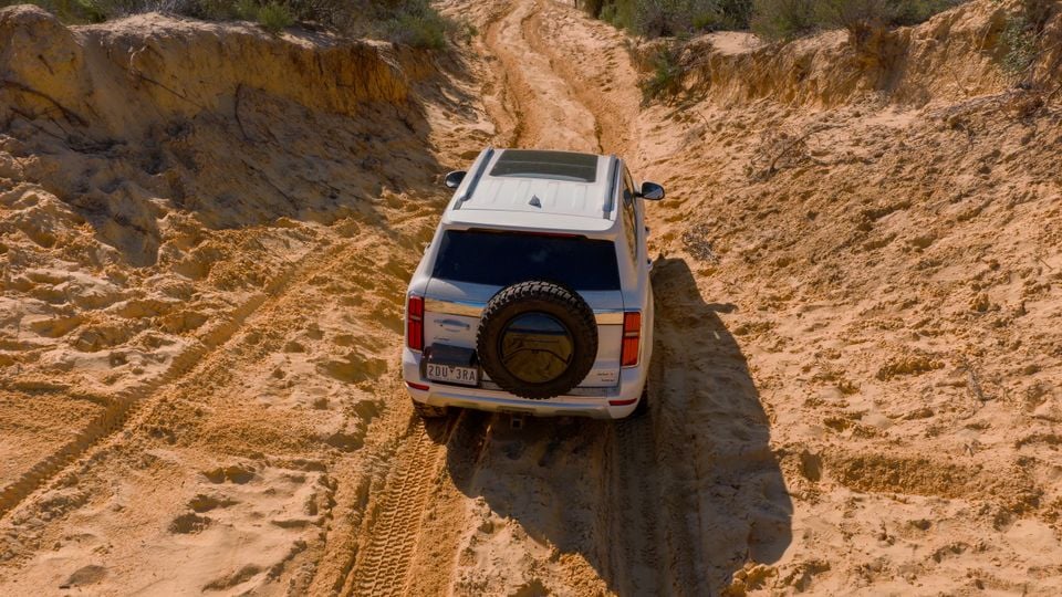 A white GWM Tank 500 SUV seen from above and behind, navigating a sandy off-road track through a desert landscape