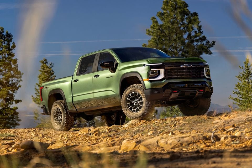 A green 2027 Chevrolet Silverado Trail Boss crew cab ute parked on a rocky hilltop among pine trees, seen from the front three-quarter angle