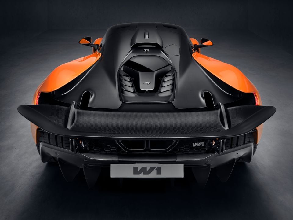 Rear view of the McLaren W1 supercar in orange and carbon black, showing its large rear wing, central exhaust outlets and exposed engine bay, photographed in a studio setting.