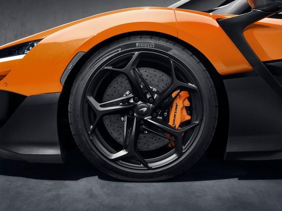 Close-up of a McLaren W1 front wheel showing a dark multi-spoke alloy wheel with Pirelli tyre, carbon ceramic brake disc, and orange McLaren-branded brake caliper against the orange bodywork and carbon fibre bodykit.