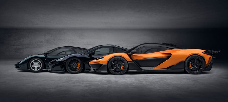 Three McLaren supercars lined up side by side in a studio, showing the evolution from the F1 (dark green) to the P1 (black) and the new W1 (orange and black).