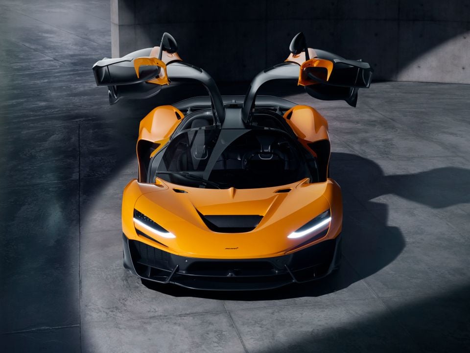 An orange McLaren W1 supercar seen from the front with both dihedral doors open wide, photographed indoors on a concrete floor.