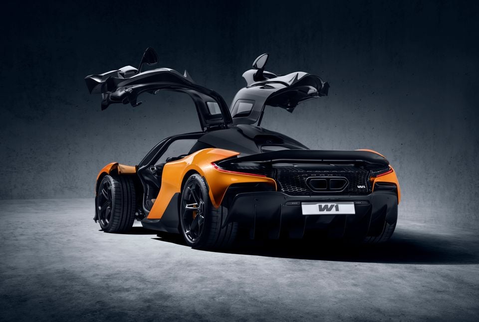 A McLaren W1 supercar in orange and black with dihedral doors open wide, seen from the rear three-quarter angle in a studio setting