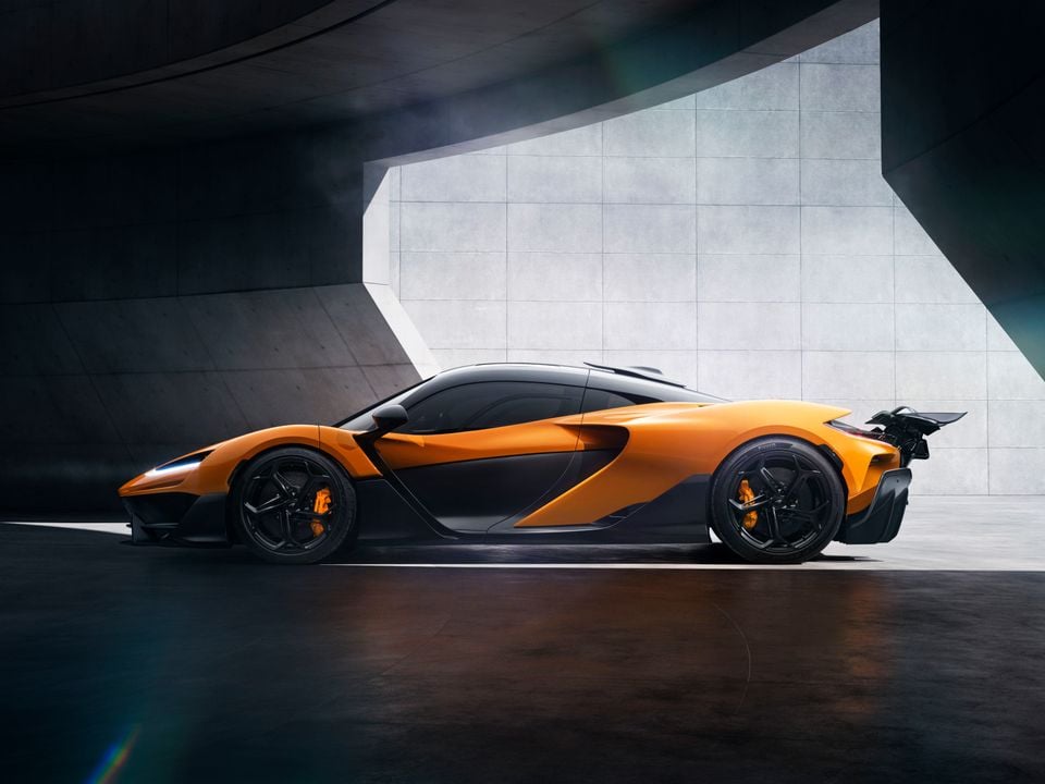 An orange McLaren W1 supercar seen from the side, featuring aggressive aerodynamic bodywork and a large rear wing, set against a dramatic concrete backdrop.
