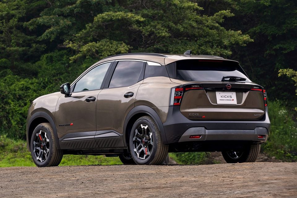 A bronze Nissan Kicks Rock Creek SUV seen from the rear three-quarter angle on a gravel surface with greenery in the background