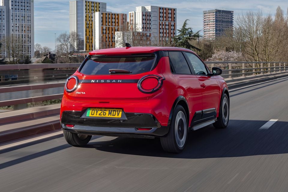 A red Nissan Micra hatchback driving on an elevated city road, seen from the rear quarter.