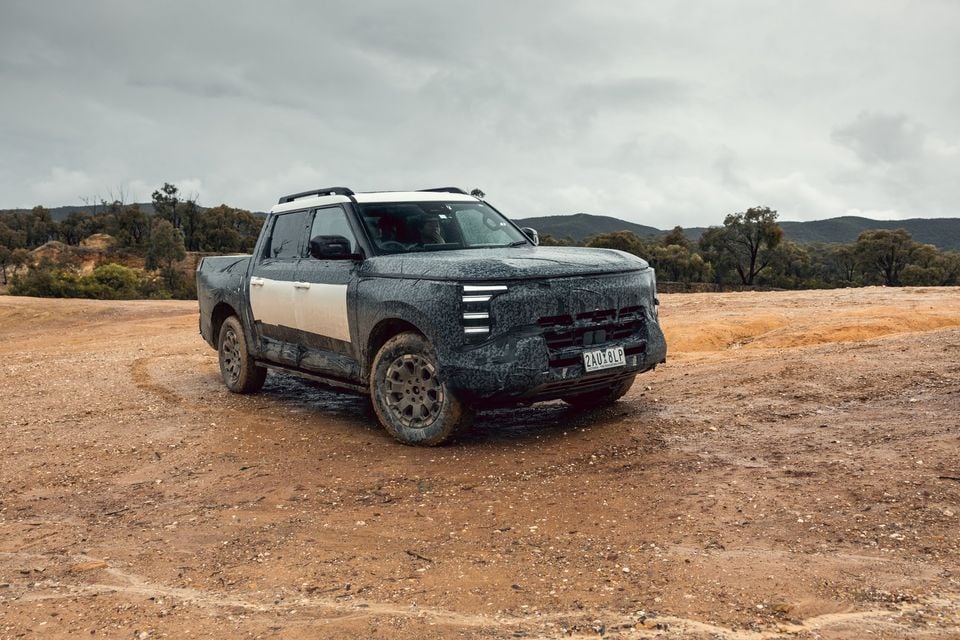 A camouflaged Nissan Navara Pro PHEV prototype dual-cab ute parked on a muddy dirt clearing under overcast skies, seen from the front