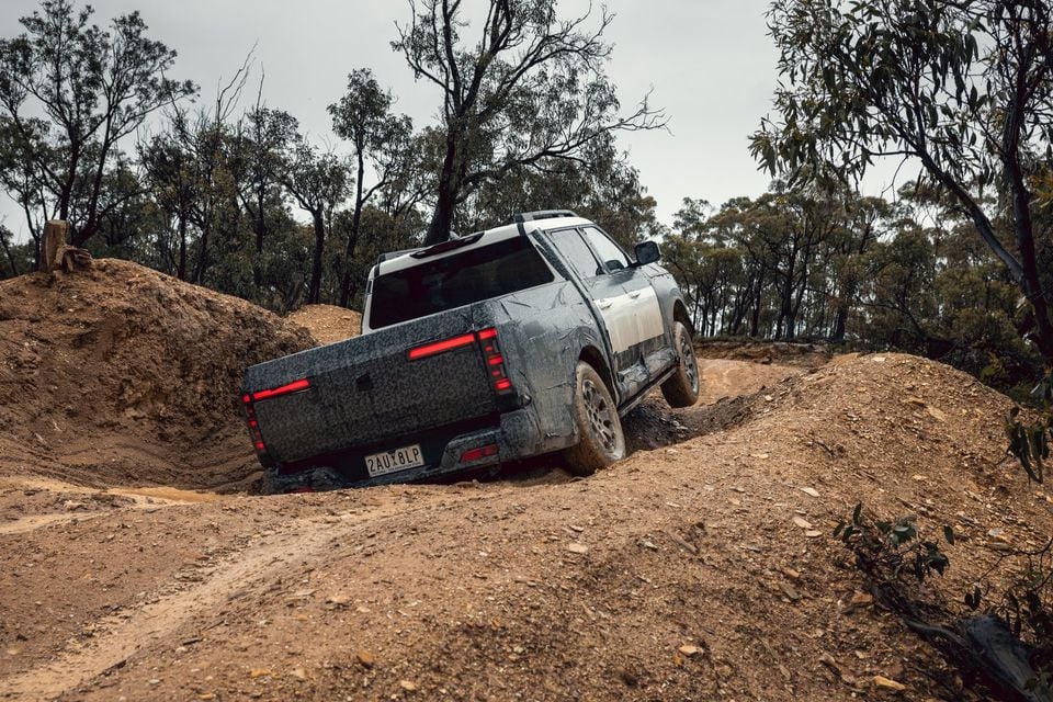 A camouflaged Nissan Navara Pro prototype dual-cab ute seen from the rear, driving over a rutted dirt track in the bush