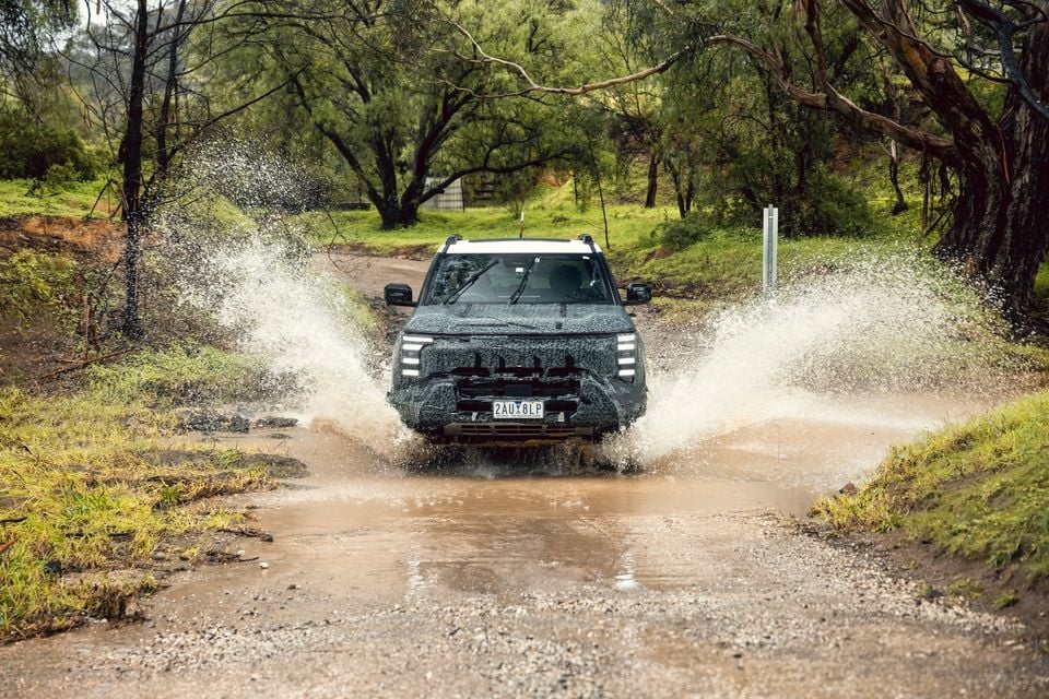A camouflaged Nissan Navara Pro prototype ute splashing through a muddy water crossing on a bush track, seen from the front