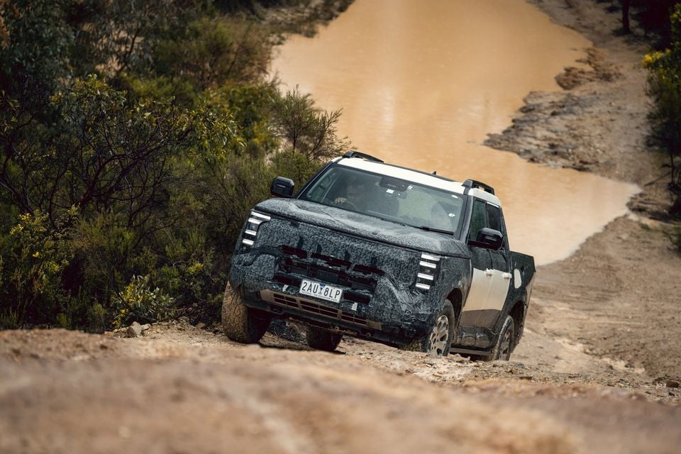 A camouflaged Nissan Navara Pro prototype dual-cab ute driving down a rocky dirt track past a muddy water crossing in the bush