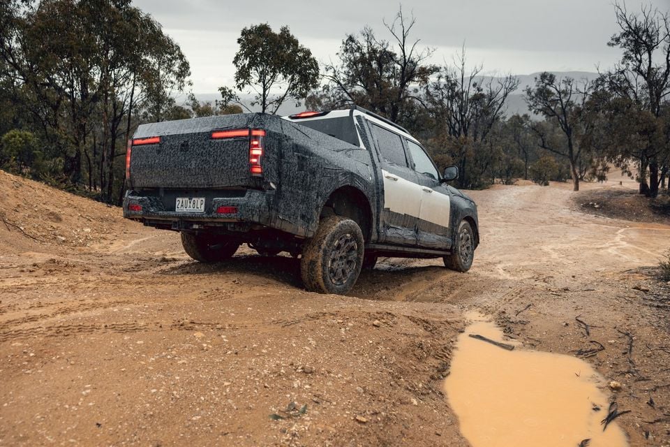 A camouflaged Nissan Navara Pro prototype dual-cab ute driving up a muddy dirt track through bushland, seen from the rear