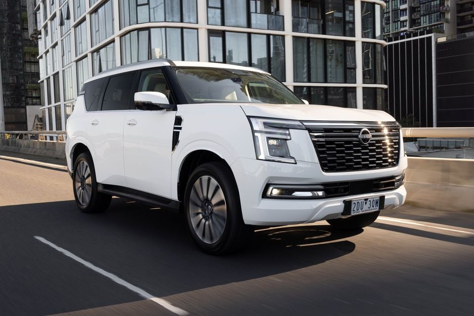 A white Nissan Patrol Y63 large SUV driving on a city road, seen from the front-side angle