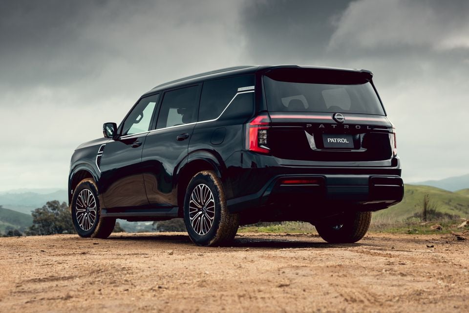A black Nissan Patrol Y63 large SUV seen from the rear three-quarter angle, parked on a dirt surface with rolling hills in the background.