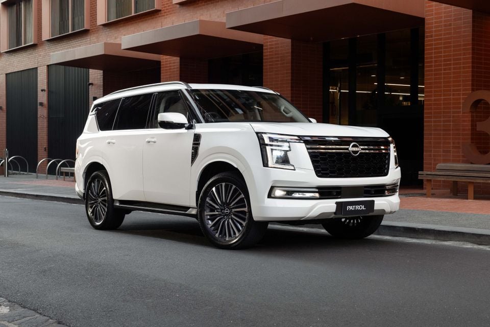 A white Nissan Patrol Y63 large SUV seen from the front-side, parked on a city street beside a brick building.