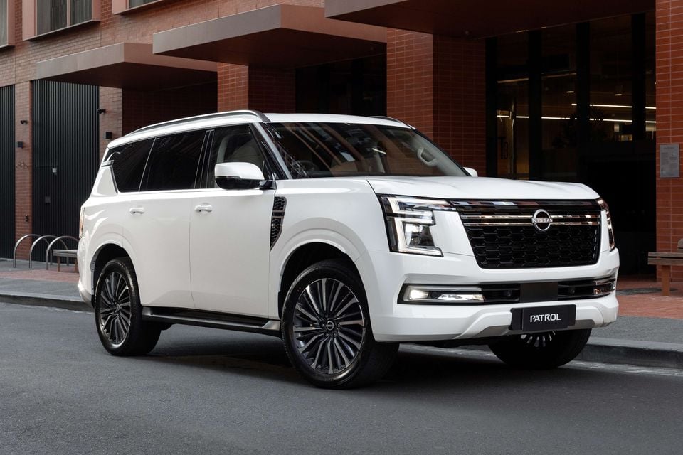 A white Nissan Patrol Y63 large SUV seen from the front-side, parked on a city street in front of a brick building