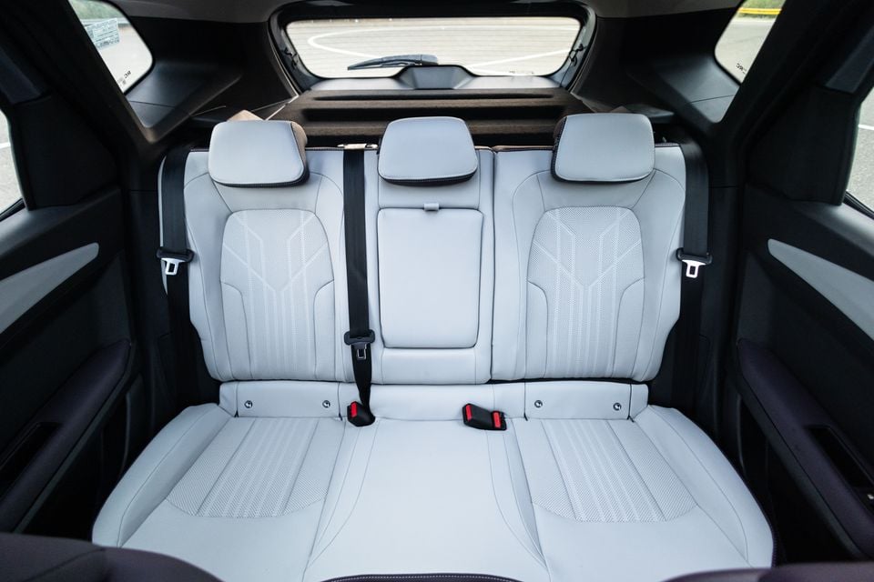 The rear bench seat of a car with light grey leather upholstery, three headrests, and seatbelts visible