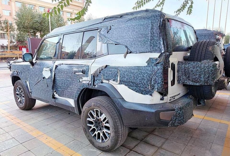 A camouflaged Nissan Terrano SUV prototype with torn disguise wrapping, seen from the rear three-quarter angle on a paved street