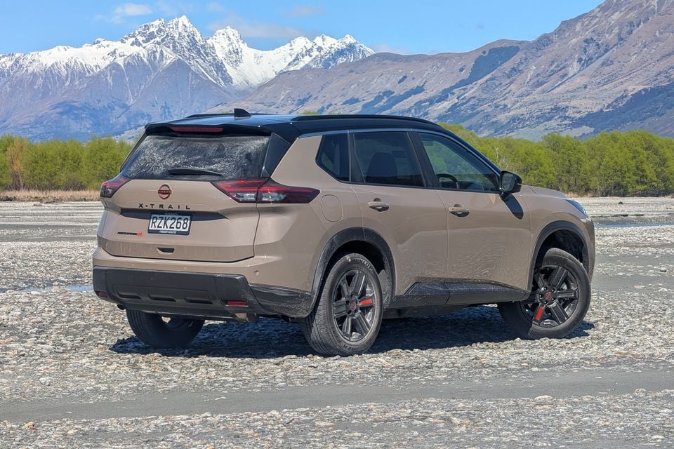A brown Nissan X-Trail SUV parked on a gravel riverbed with snow-capped mountains in the background, viewed from the rear quarter