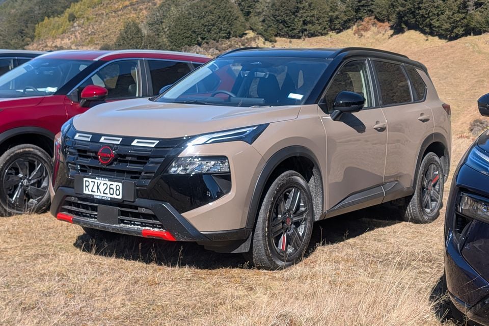 A beige 2024 Nissan X-Trail Rock Creek SUV parked on grass, seen from the front quarter view with other vehicles alongside.