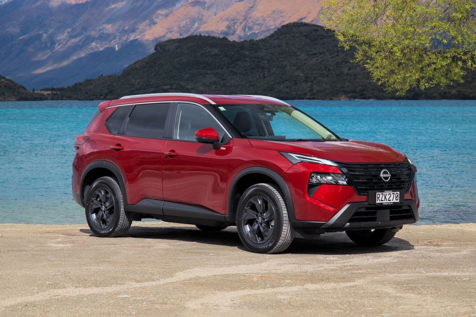 A red Nissan X-Trail ST-L e-Power SUV parked on a sandy shore beside a turquoise lake with mountains behind it.