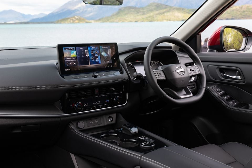 The interior of a Nissan X-Trail showing the steering wheel, dashboard and infotainment touchscreen with a lake and mountains visible outside.