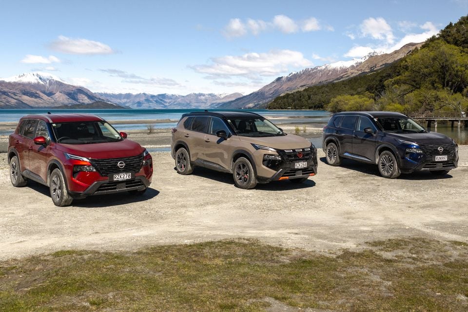 Three Nissan X-Trail SUVs in red, tan and black parked beside a lake with snow-capped mountains in the background.