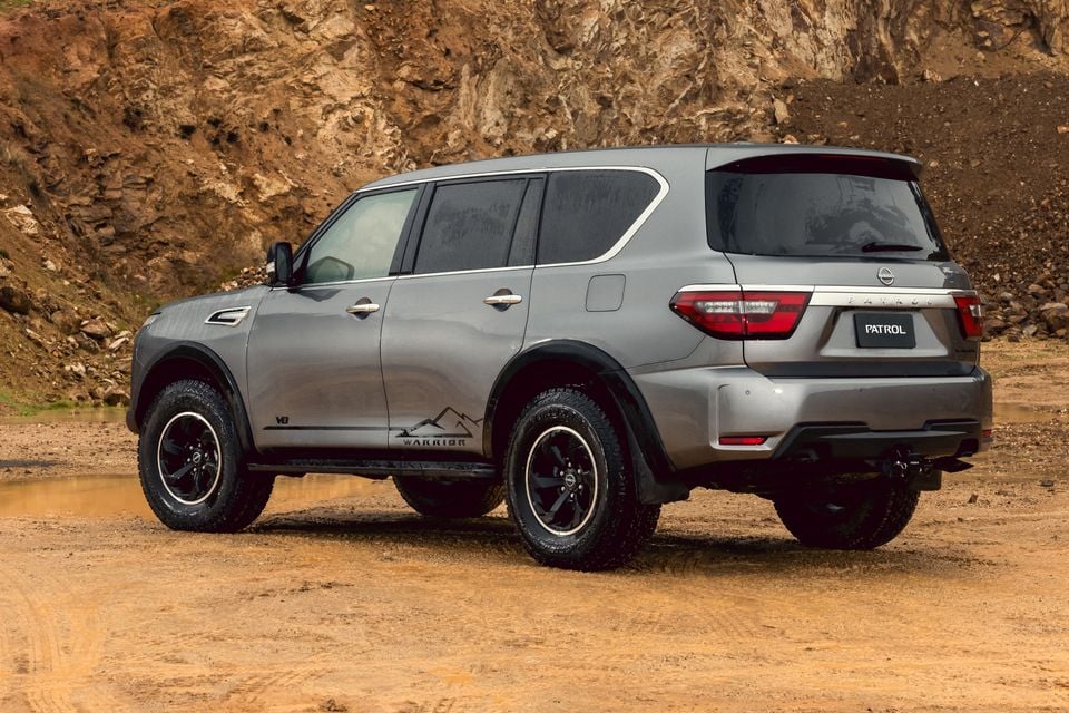 A grey Nissan Patrol Y62 Warrior SUV seen from the rear three-quarter angle, parked on dirt terrain with rocky cliffs in the background