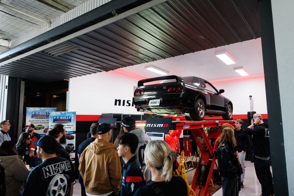 A black Nissan Skyline GT-R coupe raised on a lift, viewed from the rear, with a crowd of people watching at the NISMO Performance Centre.