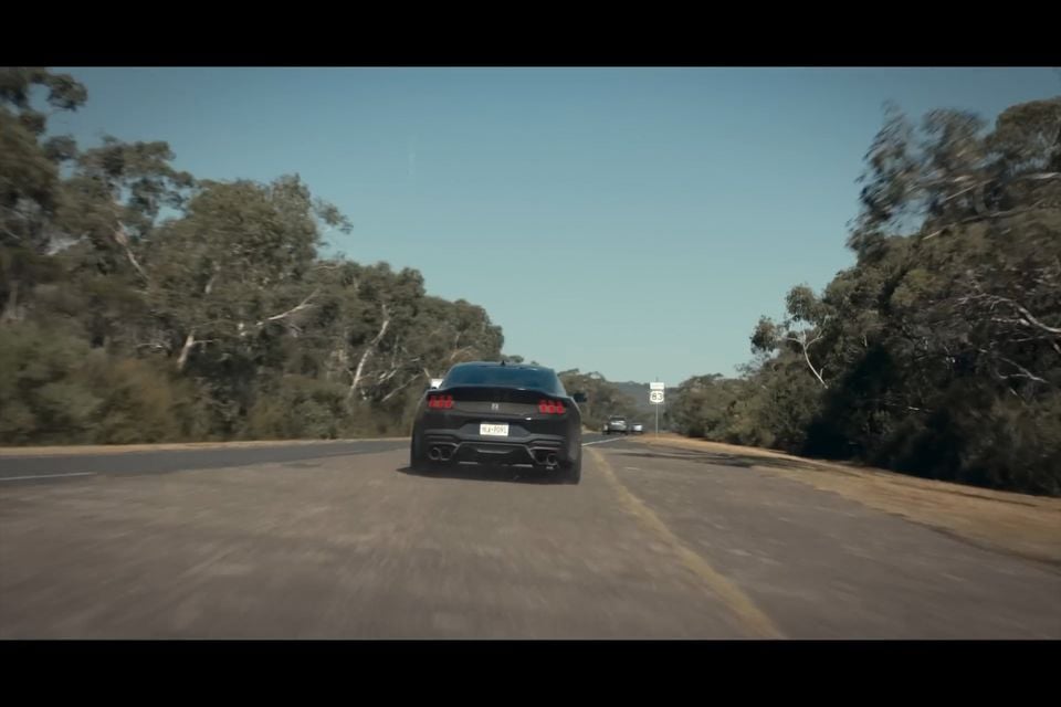 A dark-coloured sports coupe seen from the rear driving along a tree-lined country road on a sunny day.