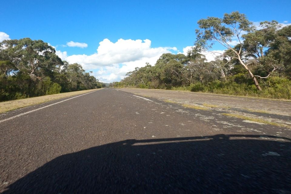 A view from a low angle along an open Australian rural road lined with eucalyptus trees under a blue sky with scattered clouds
